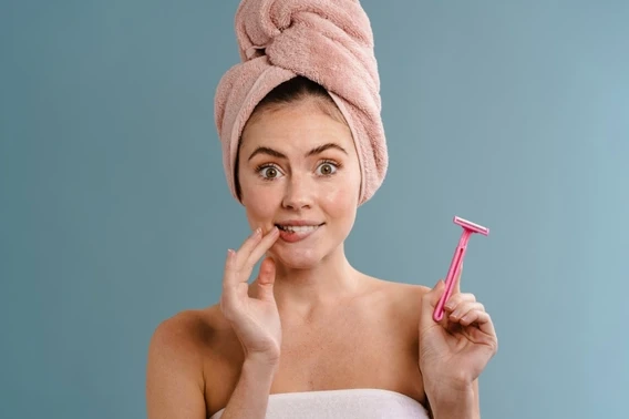 woman wrapped in a towel holding a pink razor, exploring at-home hair removal vs. professional dermaplaning benefits